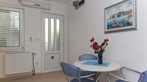 A room with a round table, four chairs, a vase with red flowers, a painting of a coastal scene, and a white door with a window.