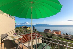 Un balcon agrémenté d’un parasol vert, de chaises en osier, d’une petite table décorée de fleurs et offrant une vue sur la mer et les montagnes.