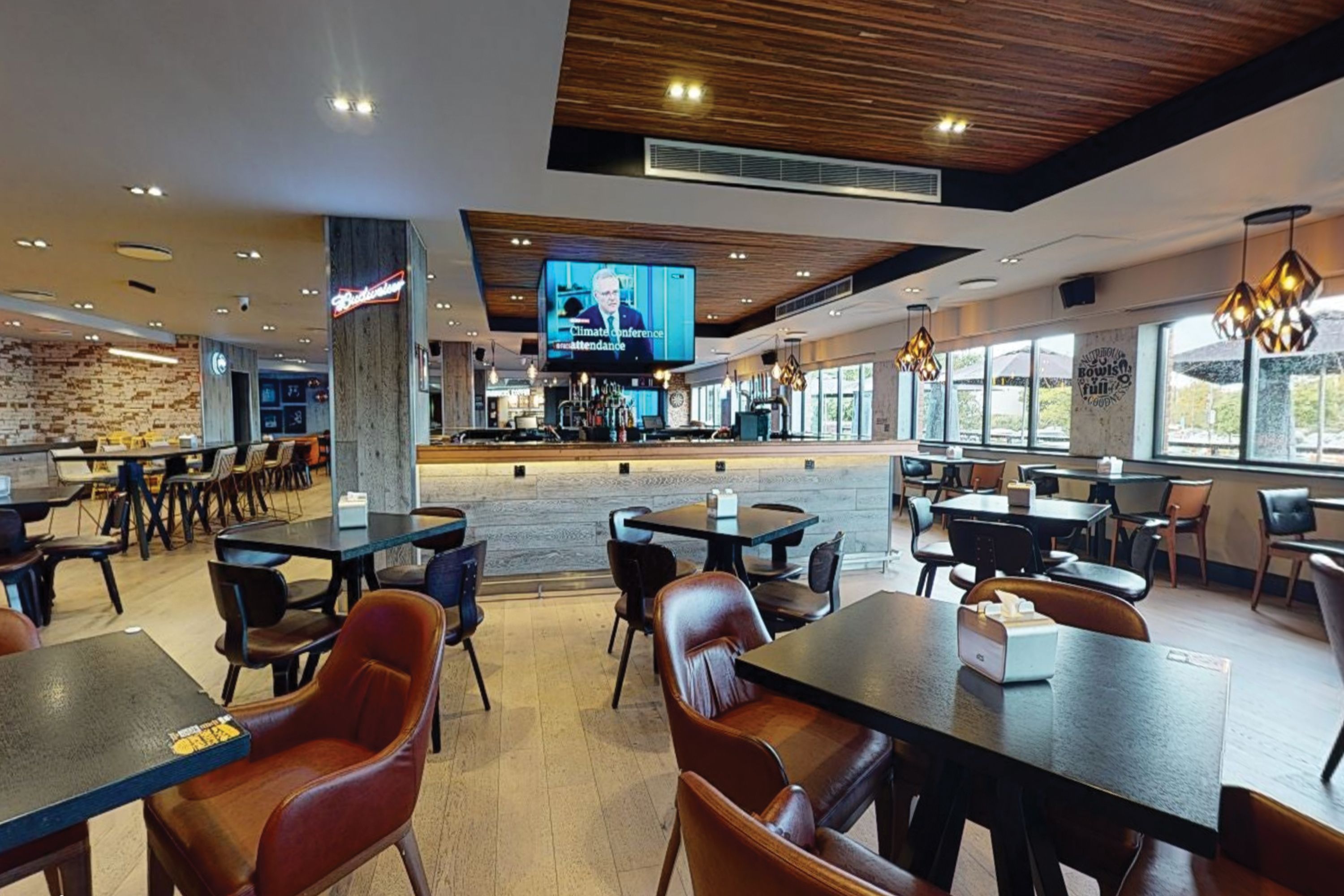 Um restaurante moderno com teto de madeira, janelas amplas e uma tela de televisão exibindo um noticiário.
