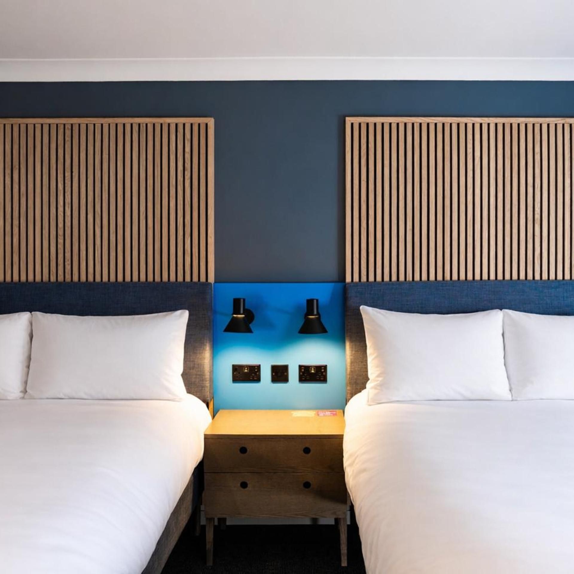 A hotel room with two beds, a nightstand, and wall-mounted lamps.