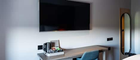A modern hotel room with a flat-screen TV mounted on the wall, a wooden desk with a chair, a small tray with items, and a mirror on the wall.