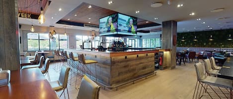 Un espace bar moderne doté d’un comptoir en bois, de nombreux sièges, notamment des tabourets de bar et des tables, ainsi que de grands téléviseurs à écran plat fixés au plafond.