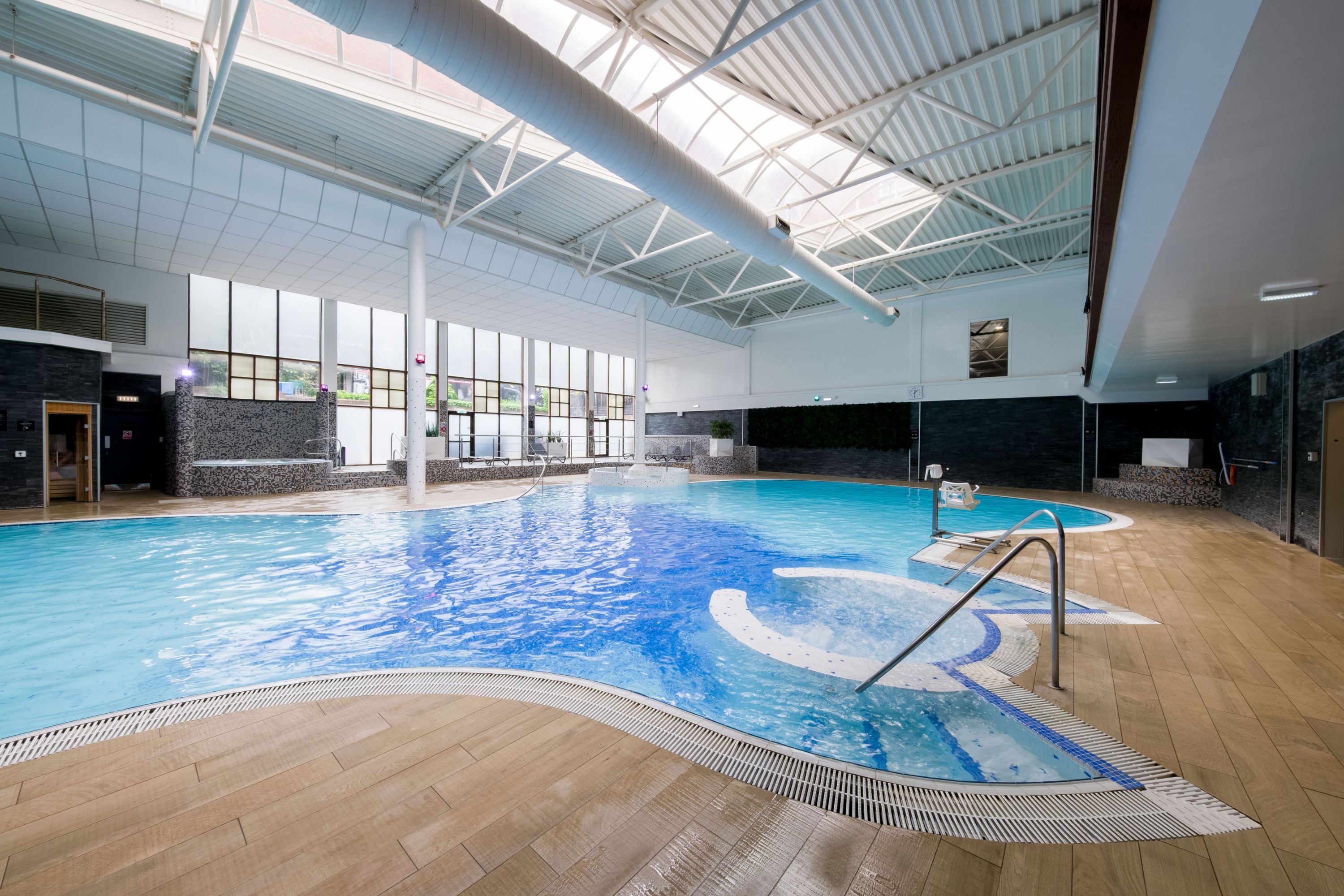 En inomhuspool med ett kaklat golv, en hoppbräda och en stor vägg med fönster.