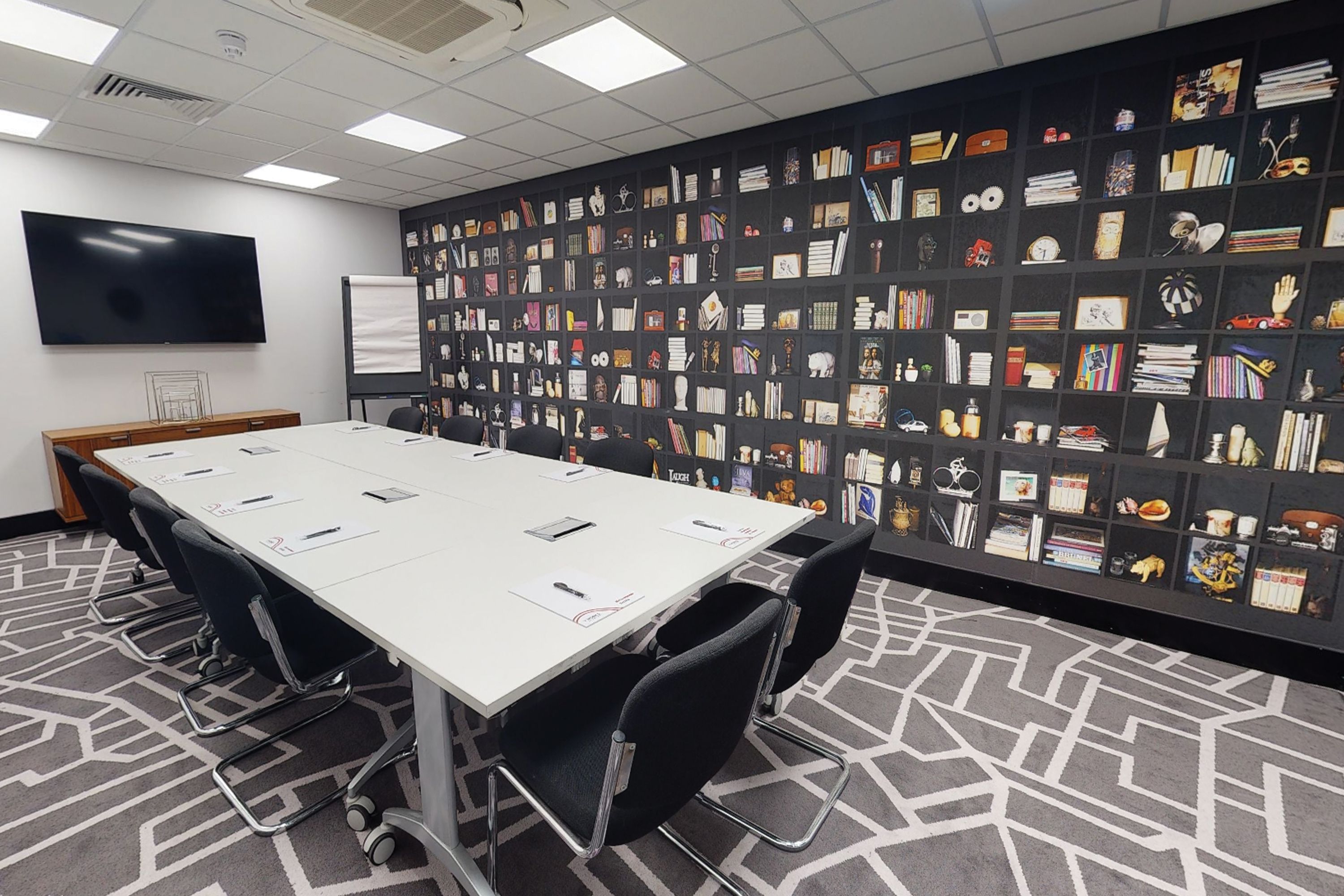 Uma sala de conferências com uma mesa comprida, cadeiras pretas, um quadro branco e uma parede decorada com diversos objetos.