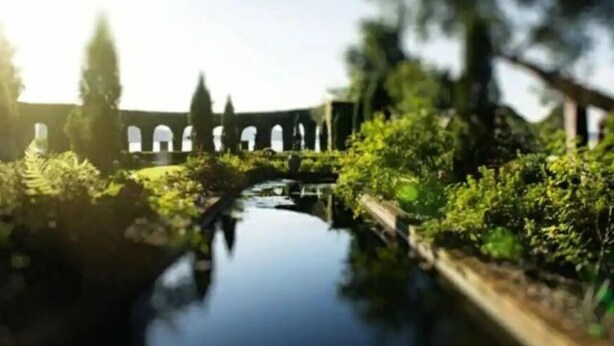 Un espacio exterior tranquilo con una fuente de agua, follaje y una estructura con arcos al fondo.