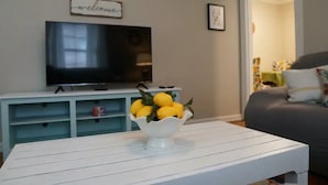 Un salón con una mesa de centro blanca, un televisor sobre un mueble, y un cuenco de limones en la mesa.