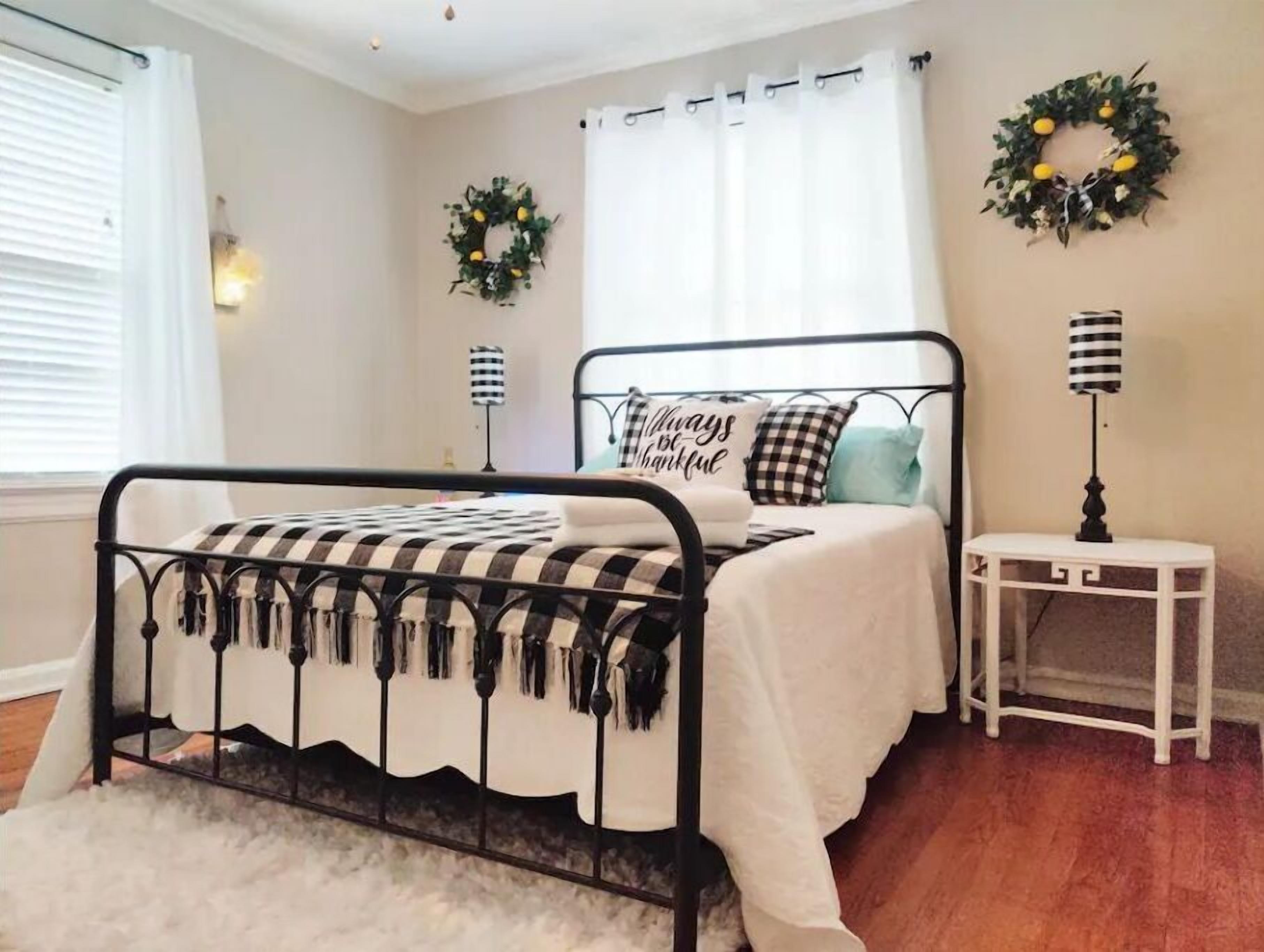 A neatly arranged bedroom with a metal bed frame, a white bedspread, and a checkered pillow. There are two wall-mounted lamps and a small white side table.