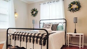Un dormitorio ordenado con estructura metálica para cama, cubrecama blanco y almohada a cuadros. Hay dos lámparas fijadas a la pared y una mesita de noche pequeña y blanca.