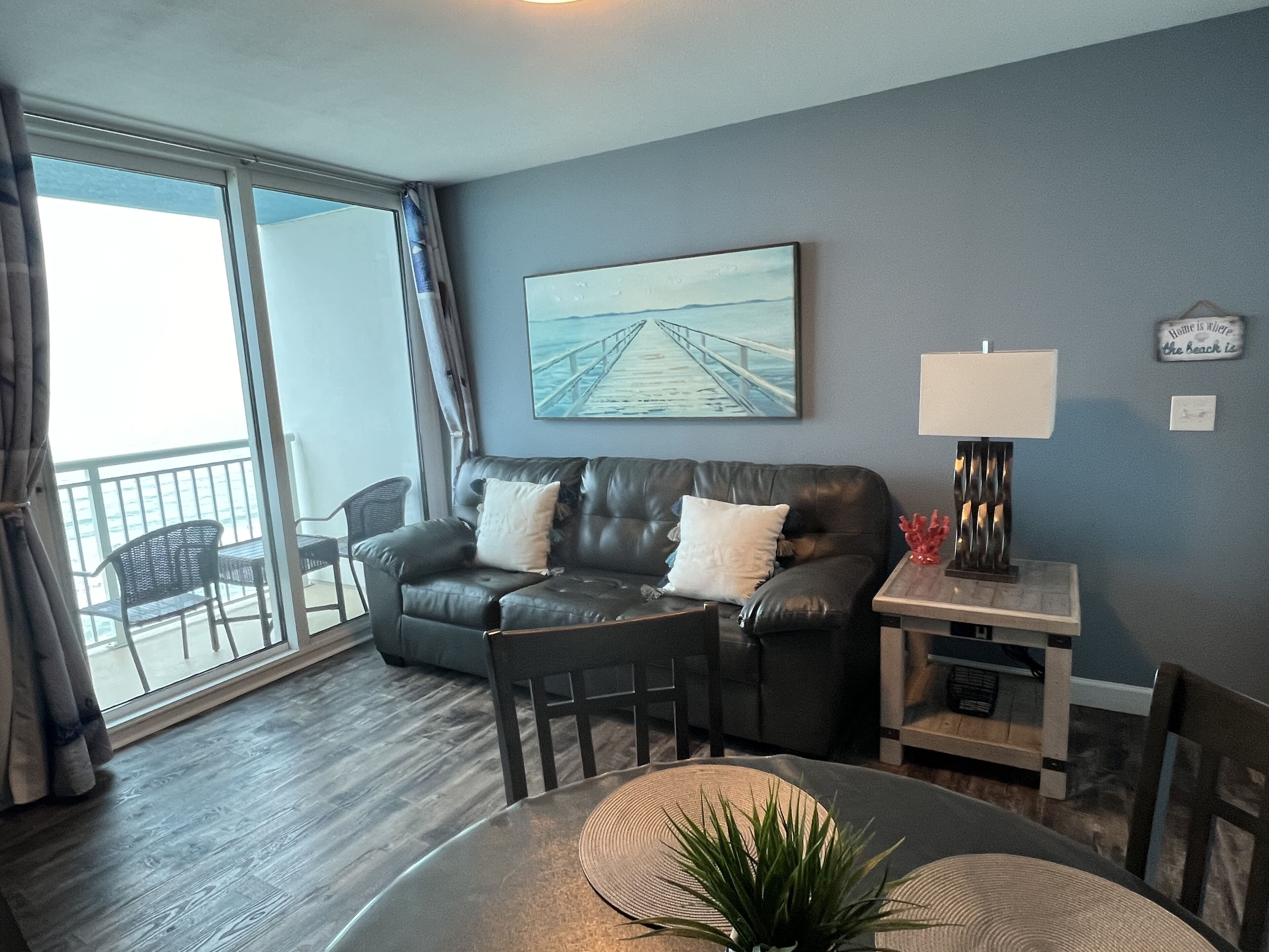 A living room with a leather sofa, a dining table with chairs, a lamp, and a view of the balcony.