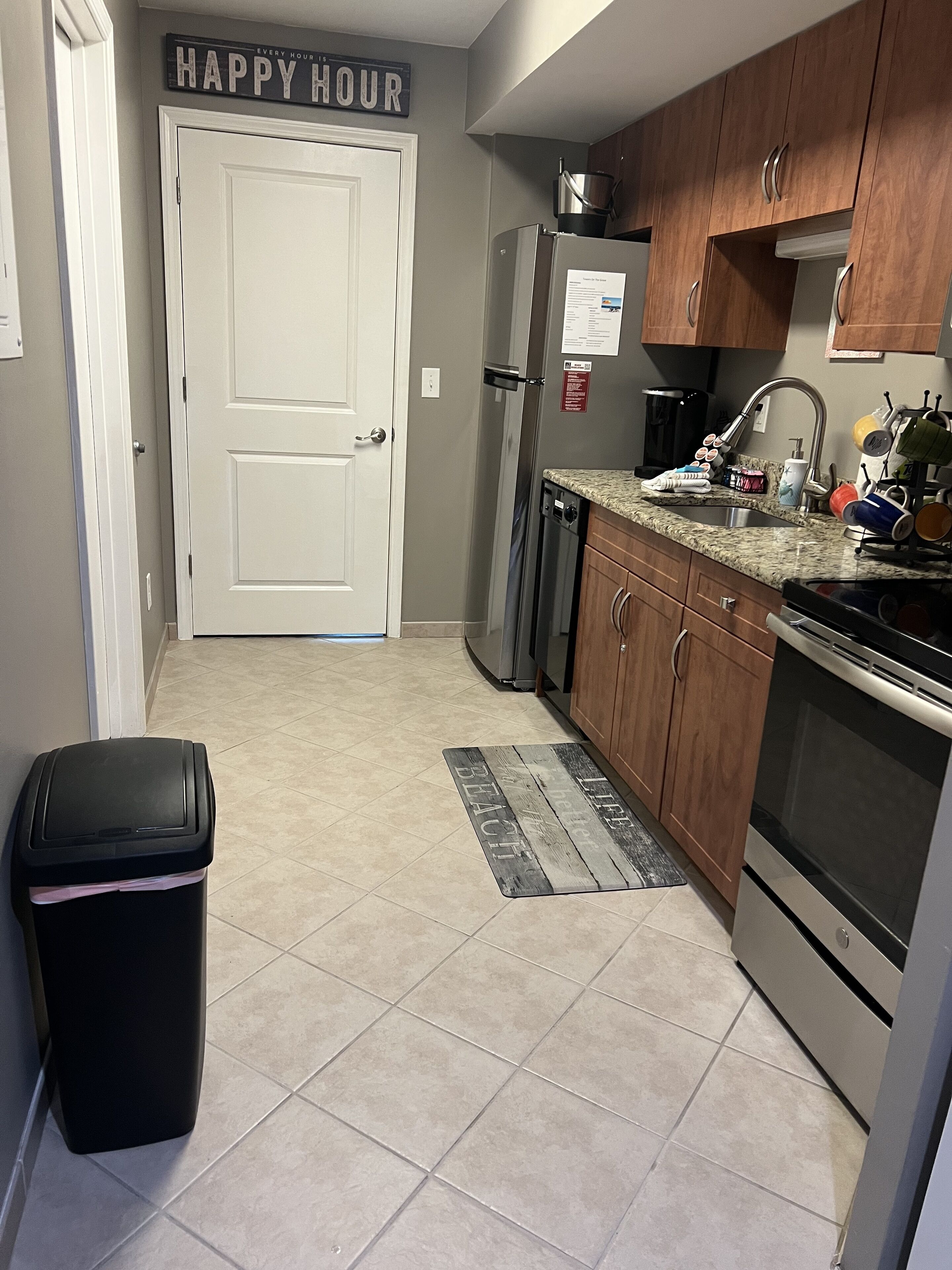 Una cocina con un refrigerador de acero inoxidable, armarios de madera, un bote de basura negro y un letrero en la pared que dice 'HAPPY HOUR'.