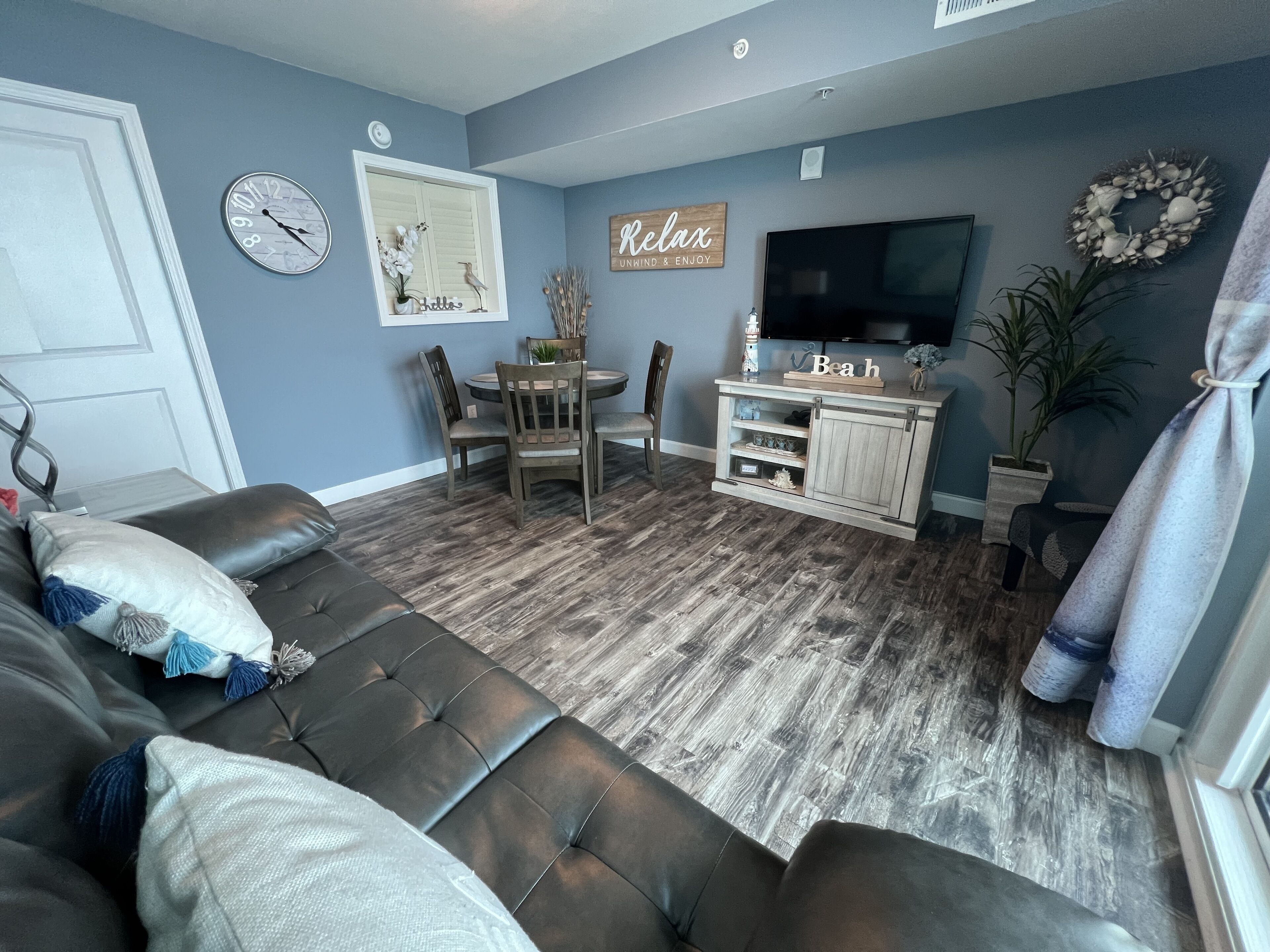 Ein Wohnzimmer mit einem dunkelbraunen Ledersofa, einem Holzesstisch mit Stühlen, einem an der Wand befestigten Fernseher und einer Kübelpflanze.