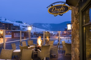 An outdoor seating area with wicker chairs and a fire pit, overlooking a snowy mountain landscape.