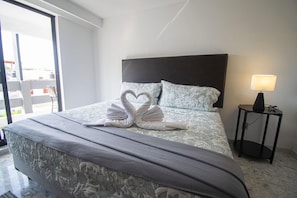 Ein ordentlich bezogenes Bett mit einem blumigen Bettüberwurf und Kissen, eine herzförmige Handtuchgestaltung, eine Nachttischlampe und Blick durch ein großes Fenster.