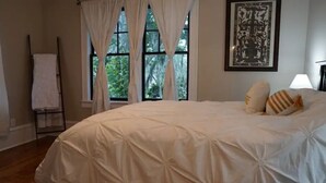 Un dormitori amb un llit gran, cortines blanques, un quadre al mur i un sòl de fusta.
