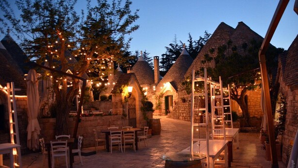 An outdoor seating area with stone tables and chairs, surrounded by stone buildings with conical roofs, under string lights. - Tenuta Monacelle (Monopoli)