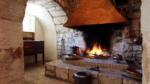 A stone fireplace with a large, ornate metal hood, a fire burning inside, and various pots and pans on the mantel. - Tenuta Monacelle (Monopoli)