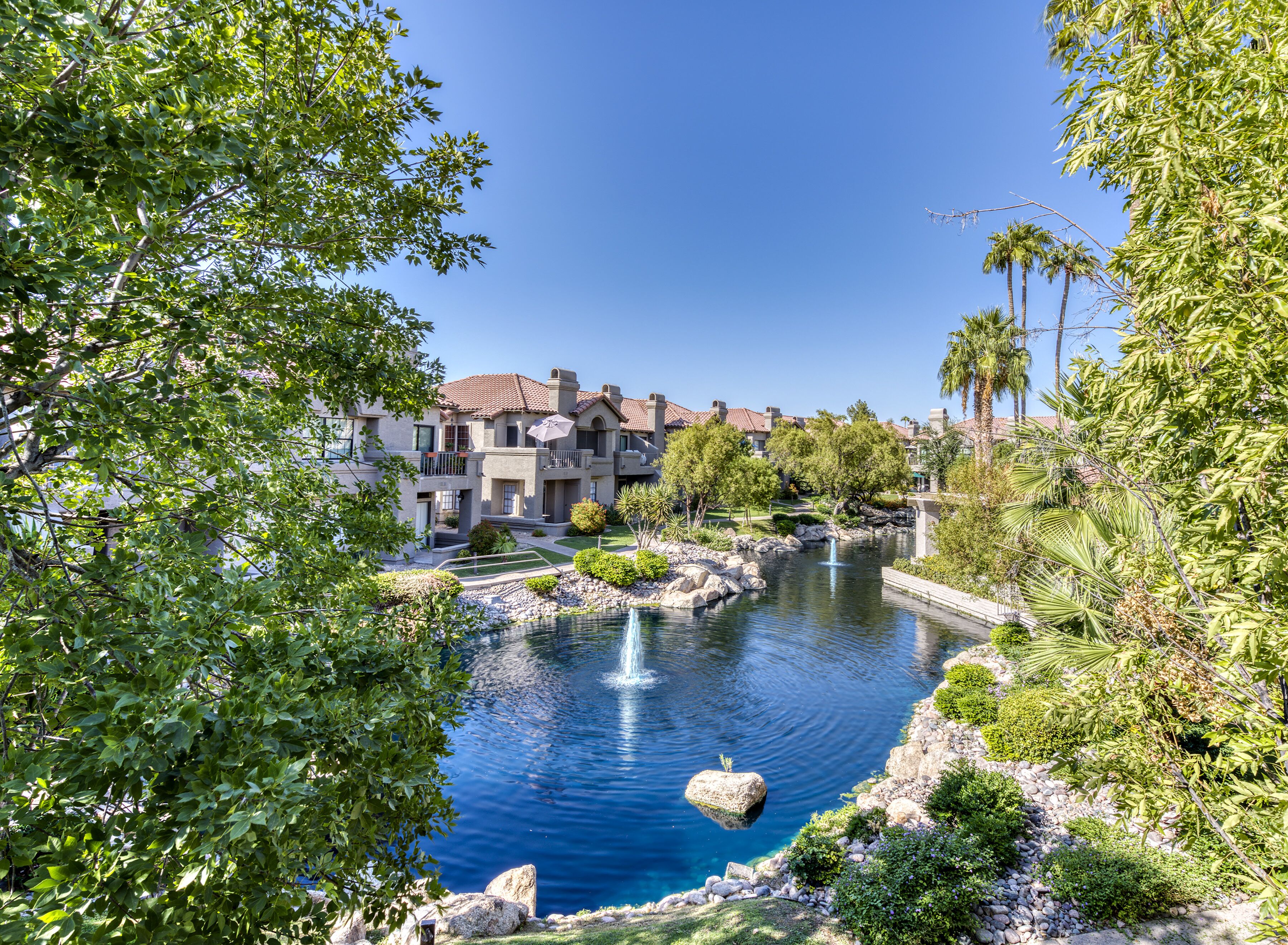 A resort with a central water feature, including a fountain, surrounded by lush greenery and palm trees.