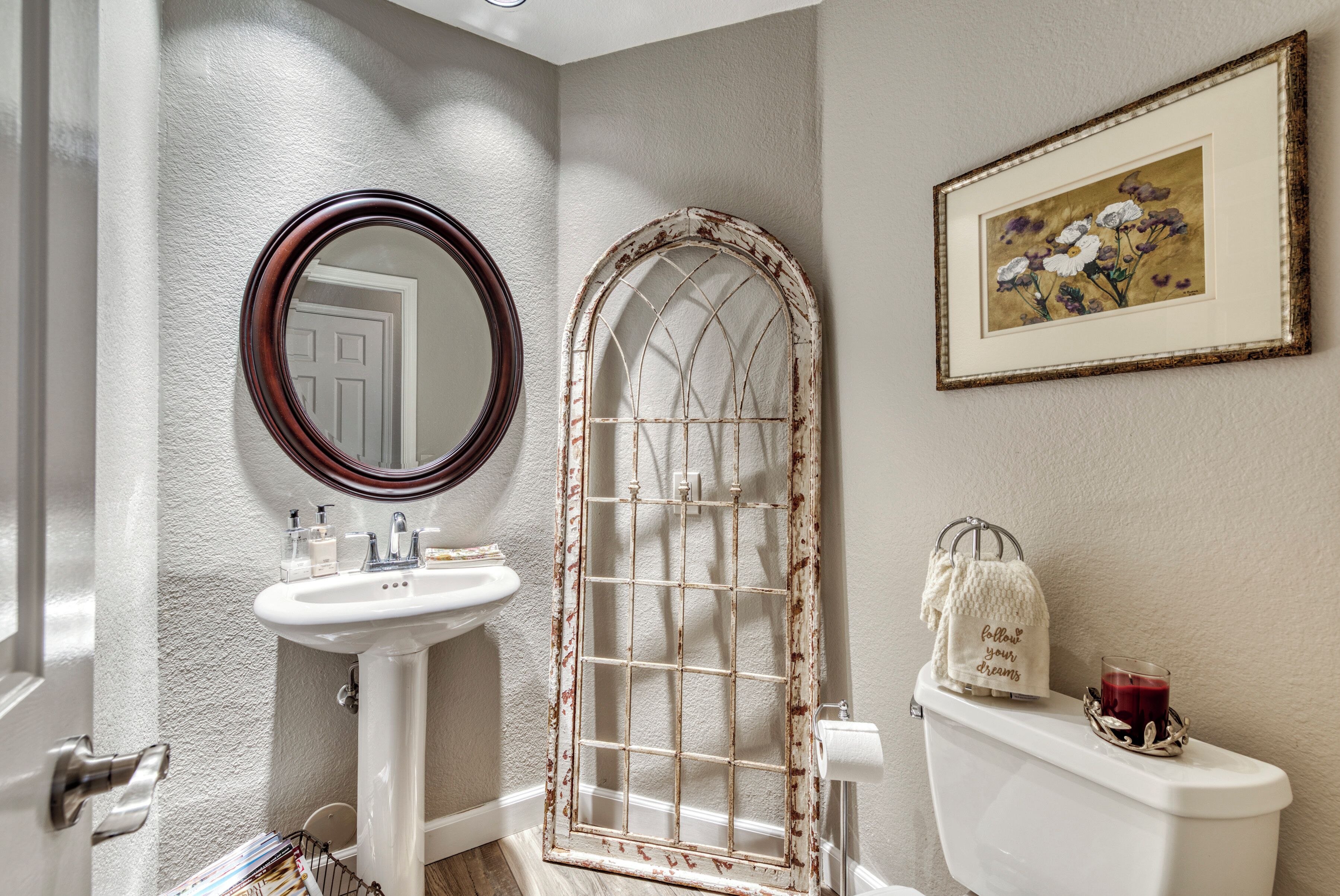 A bathroom with a white sink, a round mirror, a vintage-style window, a toilet with a candle and a towel, and a framed picture on the wall.