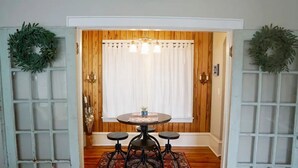 Uma sala de jantar com uma mesa redonda, três banquinhos, um tapete estampado em vermelho e uma janela com cortinas brancas.