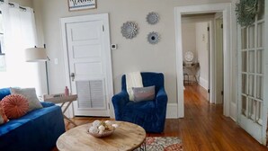Uma sala de estar com um sofá azul, uma mesa de centro redonda com objetos decorativos, uma poltrona azul e uma porta com a placa 'Welcome'.