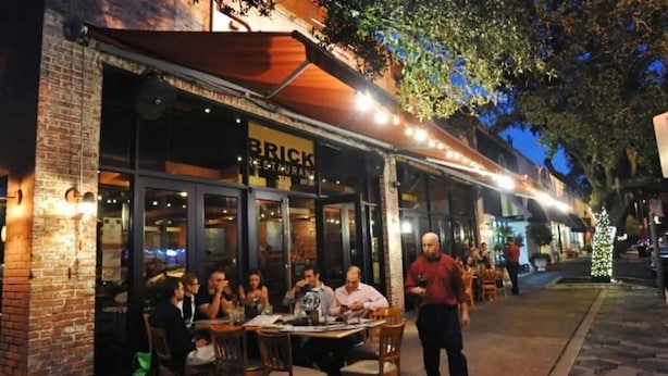 Uma área externa com mesas e cadeiras de um restaurante, com paredes de tijolos e uma placa que diz 'BRICK RESTAURANT'.