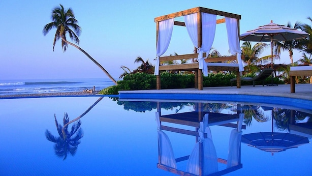 A pool with a wooden deck and a canopy structure, surrounded by palm trees and a view of the ocean. - Mishol Hotel And Beach Club (Acapulco)