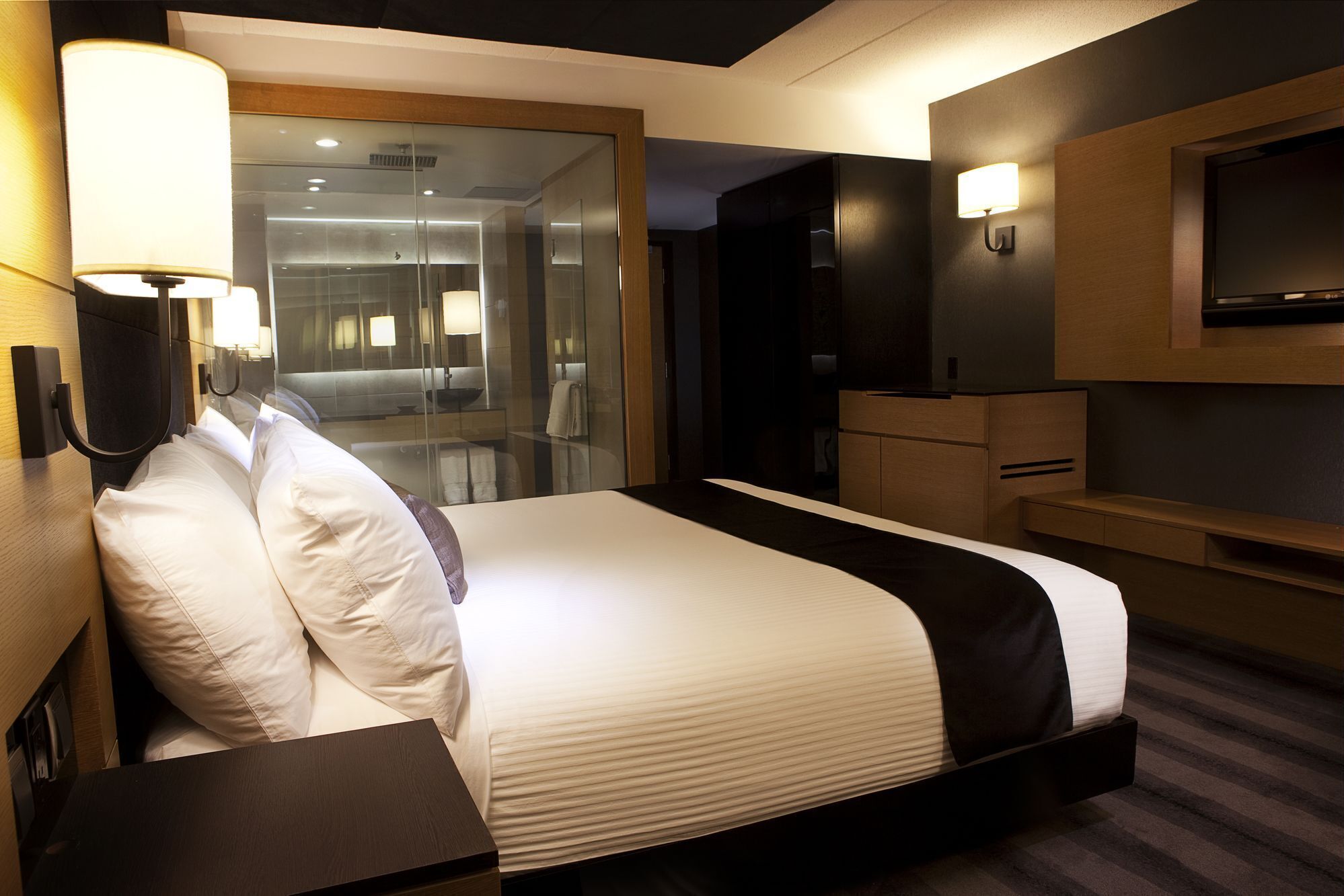 Una habitación de hotel moderna con una cama grande, una mesita de noche, un televisor y un baño con paredes de vidrio.