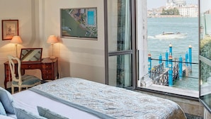 Ein Schlafzimmer mit einem großen Bett, einem Schreibtisch mit Stuhl, einem Fenster mit Blick auf ein Gewässer und Gebäude sowie einem gerahmten Bild an der Wand.