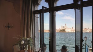 Ein Zimmer mit großem Fenster mit Blick auf ein Gewässer und ein historisches Gebäude. Ein Tisch mit Blumenvase und ein Stuhl sind ebenfalls vorhanden.