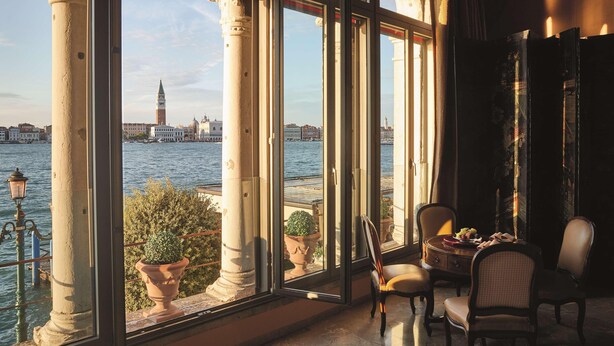 Ein Zimmer mit großen Fenstern, das auf ein Gewässer und eine ferne Stadtansicht blickt, mit einem Tisch, Stühlen und einer Topfpflanze.