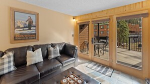 A living room with a leather sofa, a coffee table, and a painting of a skier. There is a sliding glass door leading to an outdoor deck with a table and chairs.