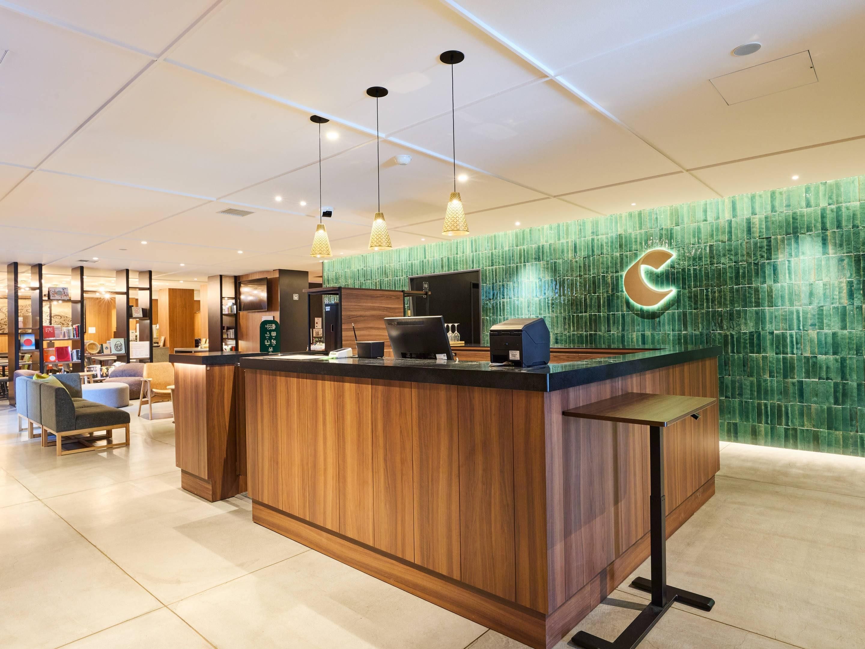Uma área de recepção de hotel com um balcão de madeira, uma parede em mosaico verde e uma área de estar com cadeiras.