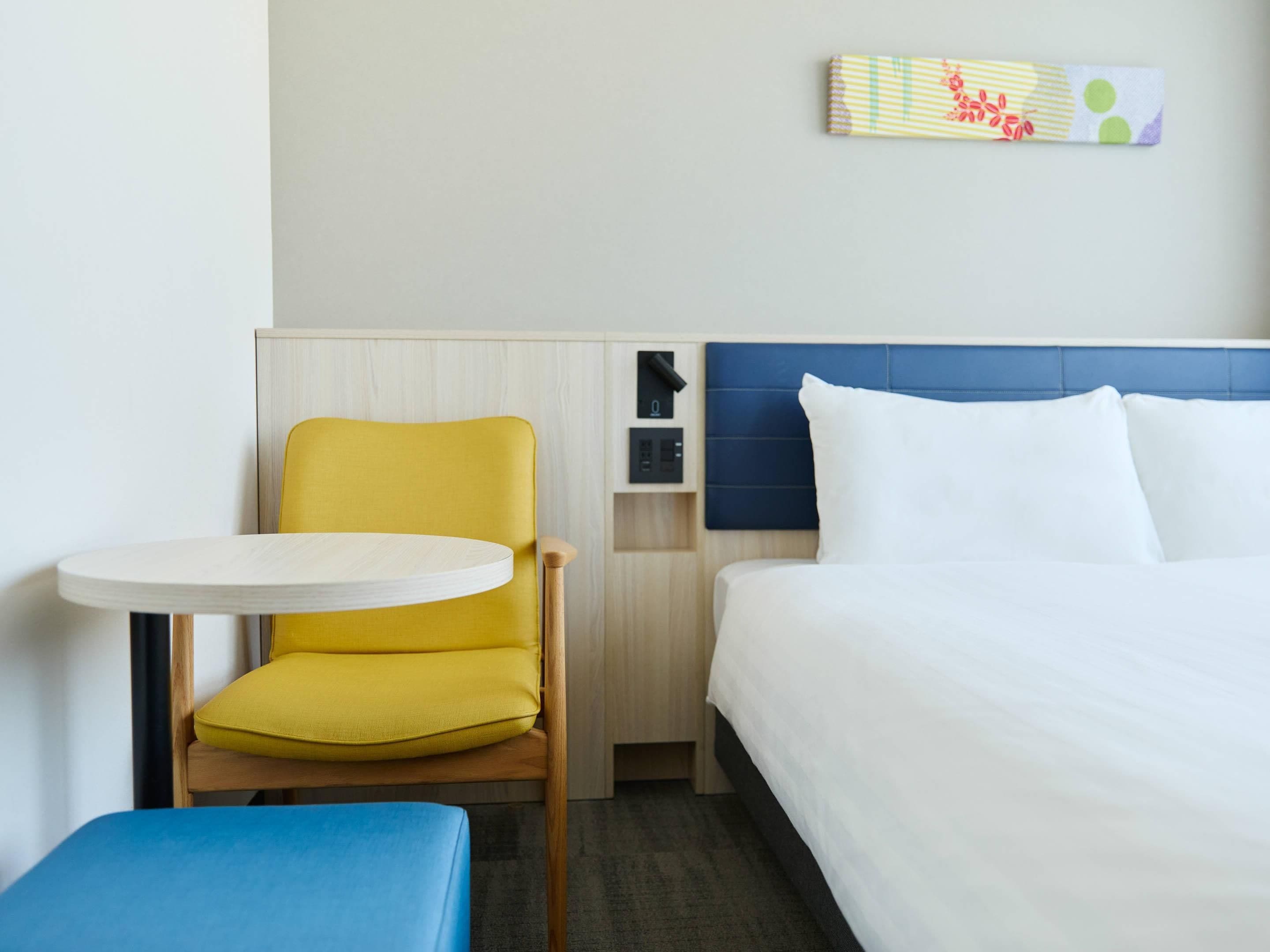 A hotel room with a bed, a small round table, a yellow chair, and a blue bench.