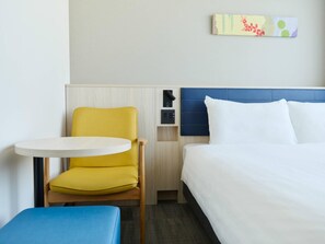 Una habitación de hotel con una cama, una pequeña mesa redonda, una silla amarilla y un banco azul.