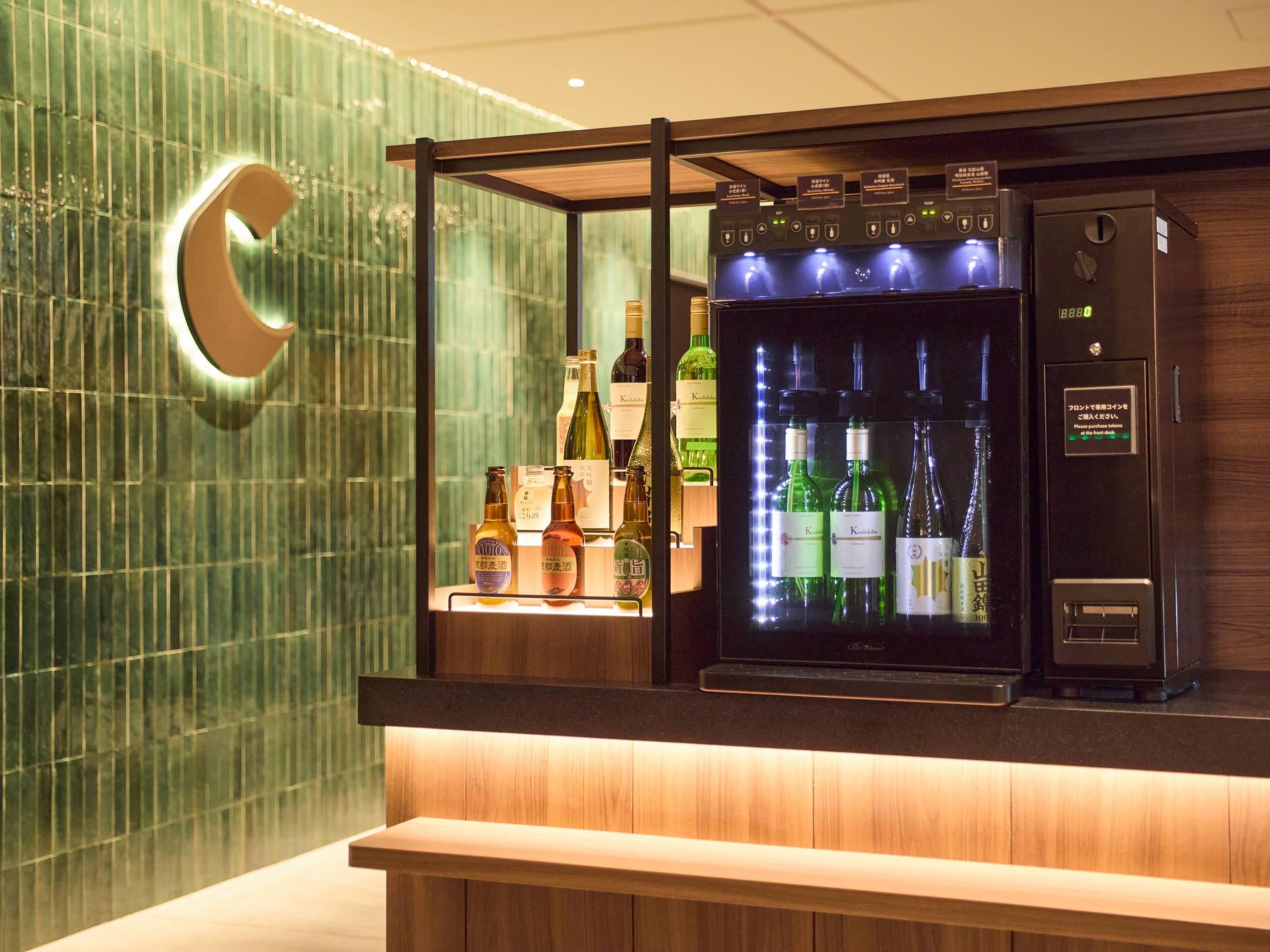 A bar area with a refrigerated display case containing various bottles, a wooden counter, and a green-tiled wall with a circular logo.