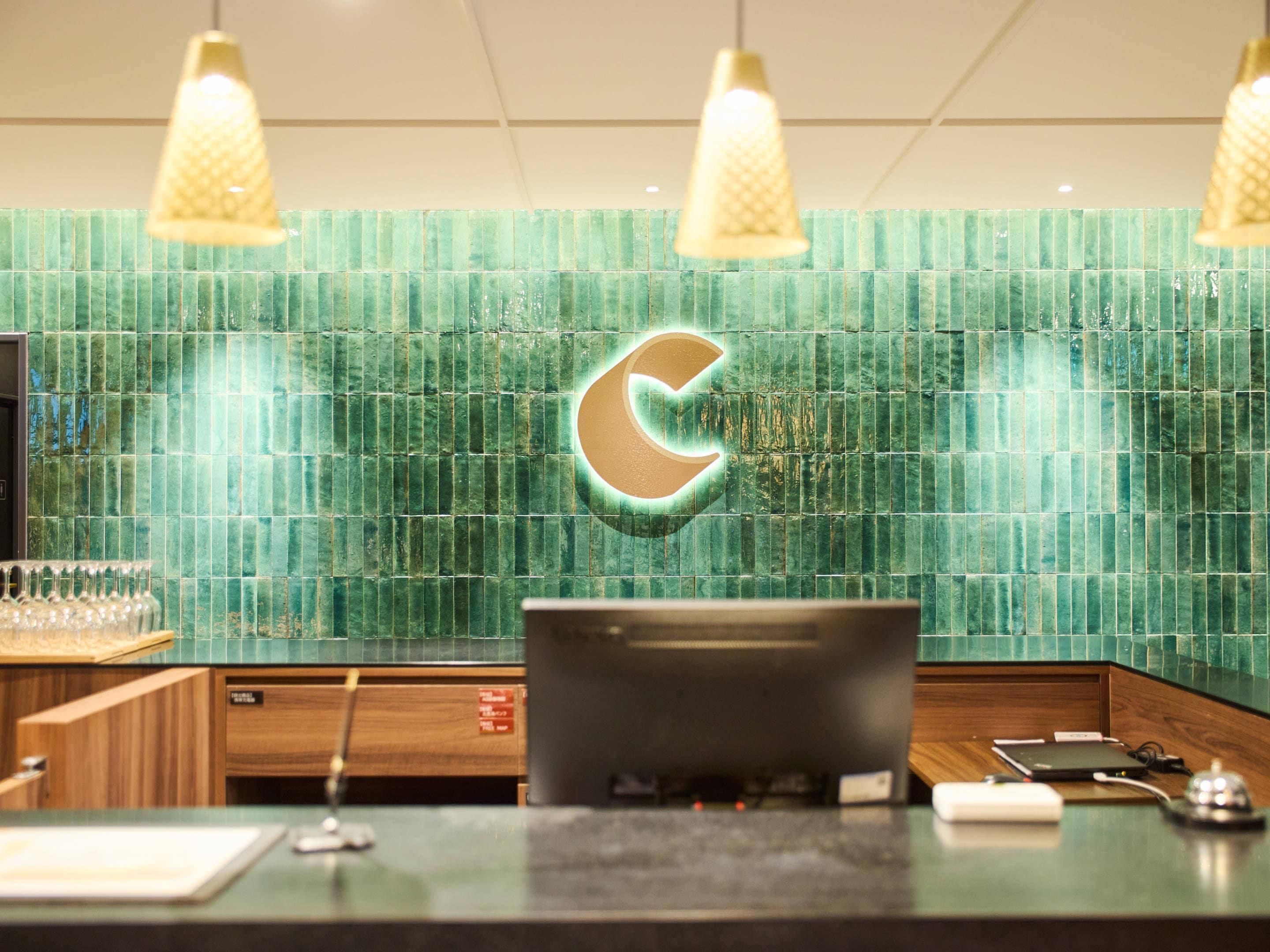 A reception desk with a green tiled wall, a large illuminated 'C' logo, and pendant lighting.