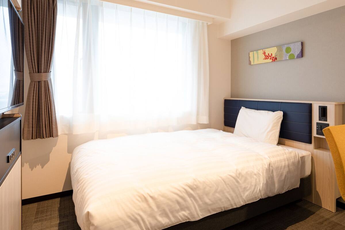 A hotel room with a bed, a desk, a chair, a TV, and a window with curtains.