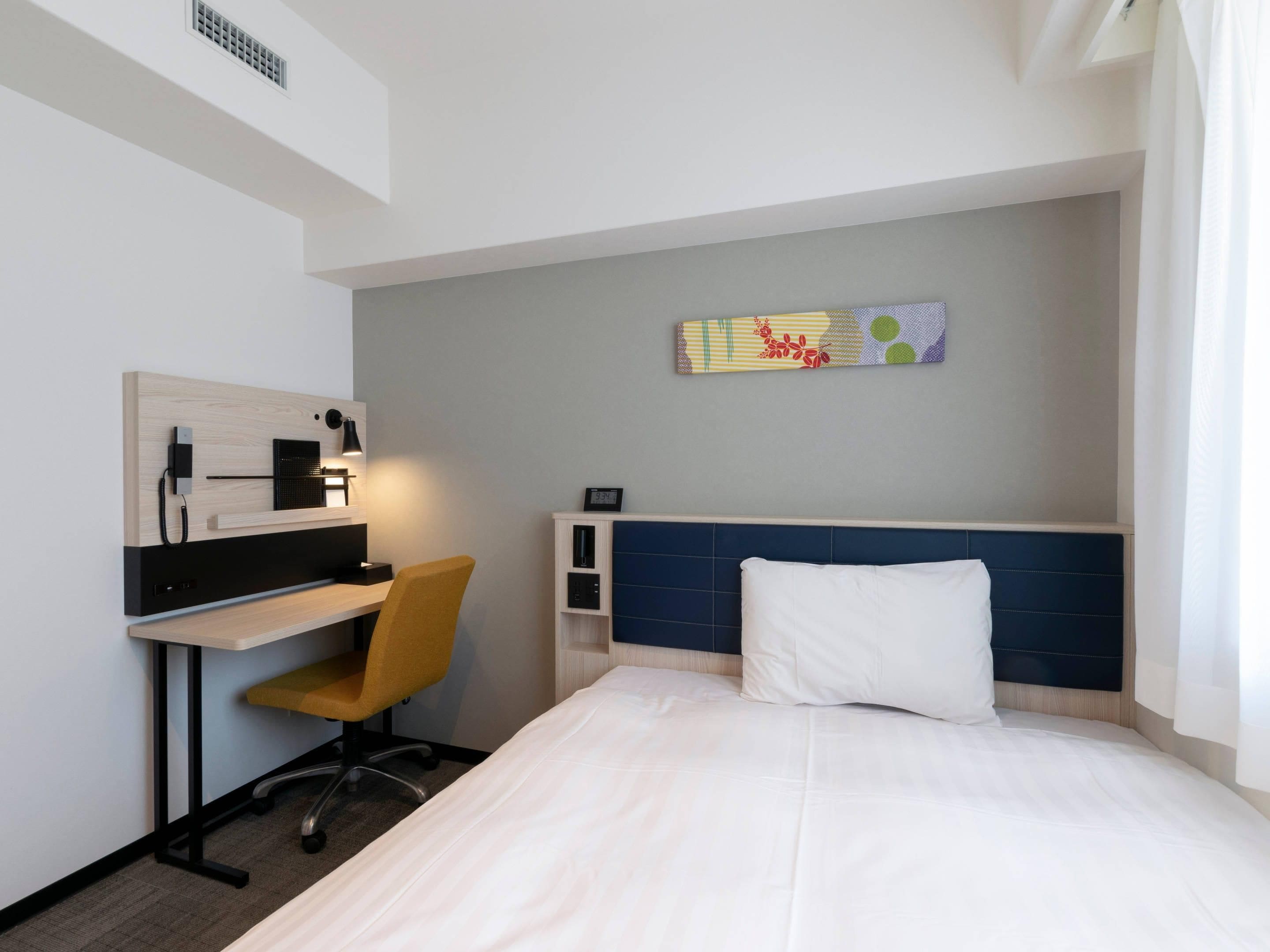 A hotel room with a bed, a desk with a chair, a wall-mounted TV, and a small shelf.