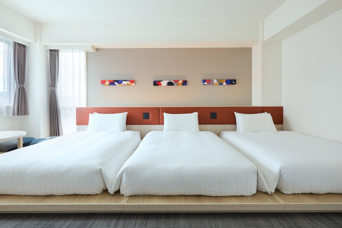 Ett hotellrum med tre enkelsängar, ett litet runt bord och en väggmonterad kontrollpanel.