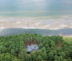 Vista aérea de uma casa grande de dois andares, rodeada por uma floresta densa, com uma praia e oceano ao fundo.