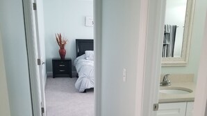 Een smalle gang die leidt naar een slaapkamer met een bed, nachtkastje en een vaas gedroogde planten.
