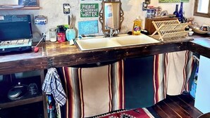 Una cocina con un lavabo, una hornilla y un estante con varias botellas y frascos. Hay un cartel que dice 'Por favor, conserve el agua'.