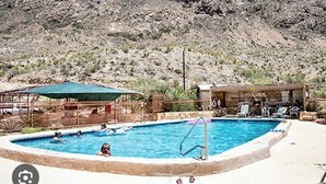 Una piscina con algunas personas, rodeada de una zona arenosa y una ladera rocosa al fondo.