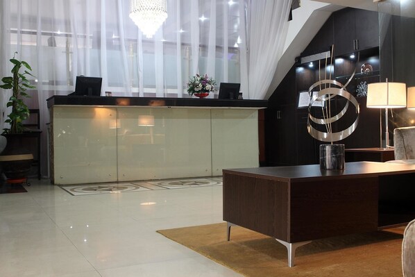 A modern hotel lobby with a reception desk, a chandelier, and a large window with sheer curtains.