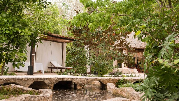 A stone bridge over a small water feature, surrounded by lush greenery and a white building with a thatched roof. - Full Hacienda Santuario Noc Ac (Mérida)