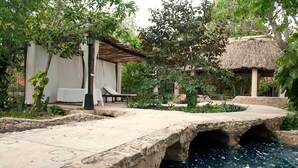 A stone bridge over a pool with a thatched-roof structure and a white building with a balcony in the background. - Full Hacienda Santuario Noc Ac (Mérida)