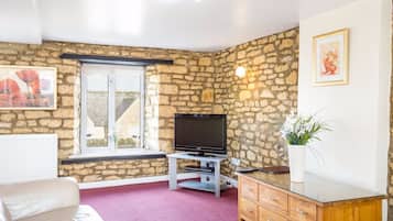 Una sala de estar con paredes de piedra, un televisor, un mueble de madera, un sofá blanco y una ventana con vistas a edificios.