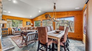 Interior espacioso de una cabaña con comedor, chimenea y grandes ventanales que ofrecen vistas del bosque circundante.