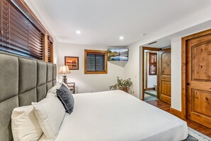 A bedroom with a large bed, wooden doors, a window with blinds, a wall-mounted TV, and a framed picture. - Tranquil 4BR on Peak 7 w/ Hot Tub, Dog Friendly! (Breckenridge)