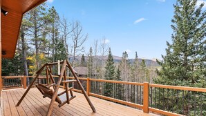 Una terraza de madera con un columpio, rodeada por una barandilla y árboles, con vistas a un paisaje montañoso.